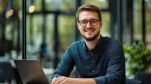Jeune apprenti souriant travaillant sur un ordinateur portable dans un espace de coworking moderne, préparant son contrat d’apprentissage avant le début de sa formation. Confiant et motivé, il profite d’un cadre lumineux et professionnel.