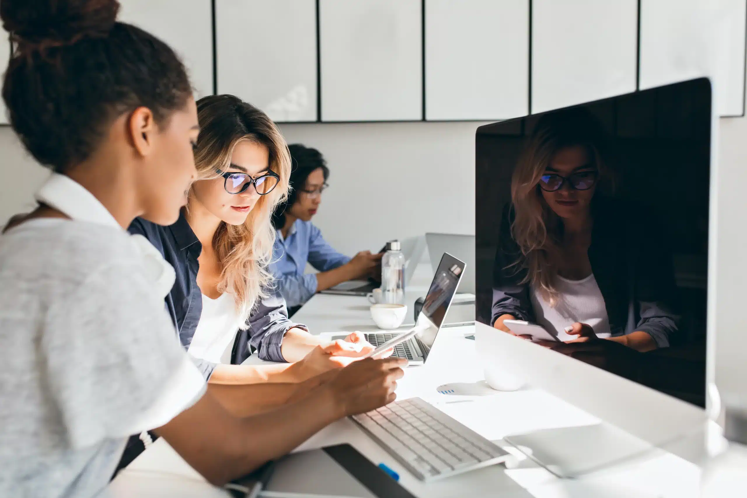 Deux professionnelles en formation SAP collaborant sur un ordinateur portable dans un environnement de travail moderne, avec d'autres participants en arrière-plan.