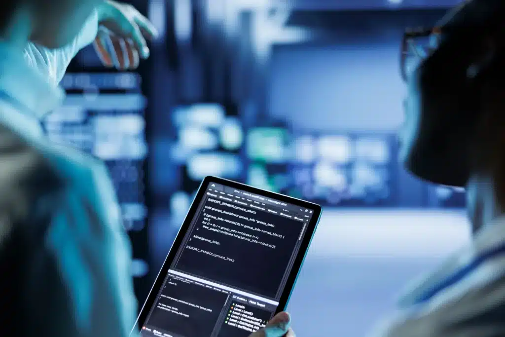 Deux professionnels de la cybersécurité analysent un code source sur tablette dans un data center, illustrant une situation typique de gestion des menaces et de compétences techniques liées à la certification CompTIA Security+.