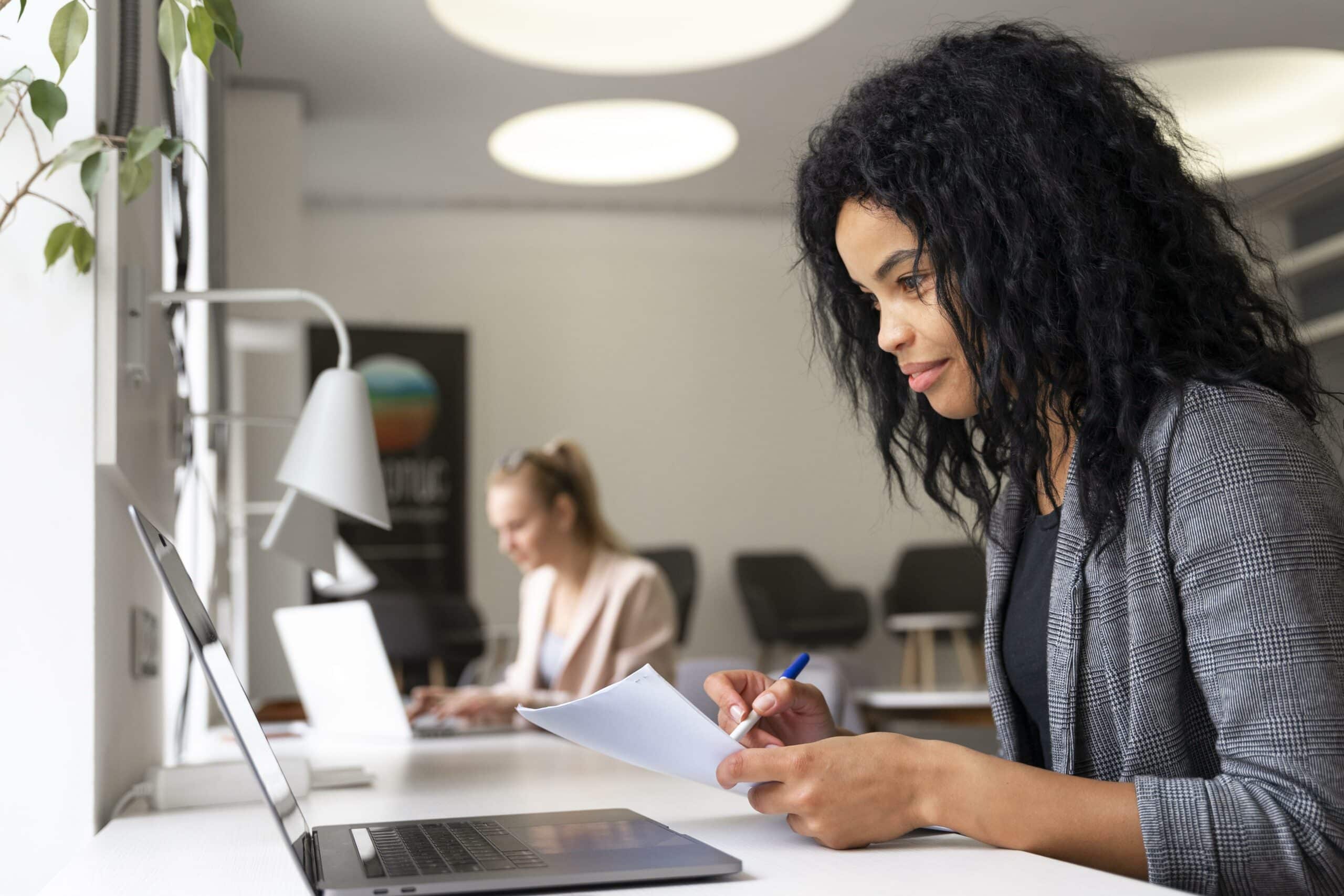 La meilleure développeuse fullstack en train de prendre des notes tout en travaillant sur son ordinateur portable dans un espace de coworking lumineux.